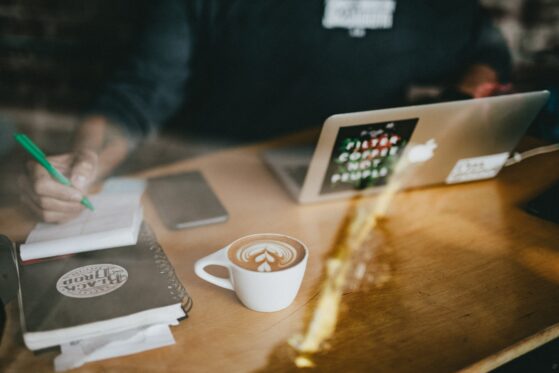 nathan-dumlao-520251-unsplash The photo shows a light wooden desk with a macbook air, cappuccino with latte art leave, notebook and a hand of a person writing