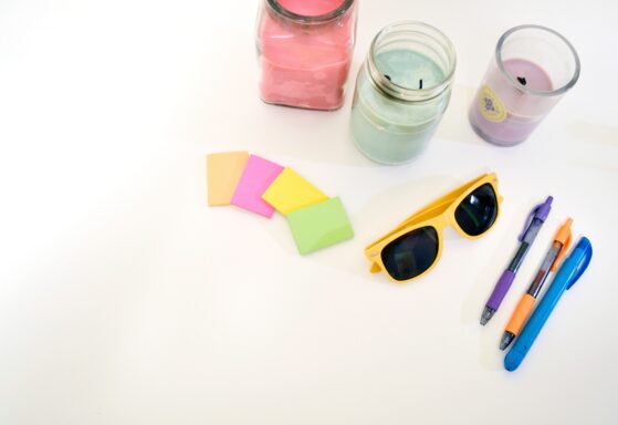 Colorful desk flat lay photo, with colorful candels in mint lavender and pink colors, yellow sunglasses, stickynotes in bright green, yellow, pink and orange, and 3 ballpoints in pin orange and blue