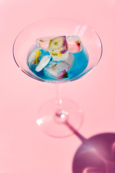 coktail glass with blue fluid and ice cubes on a pink background